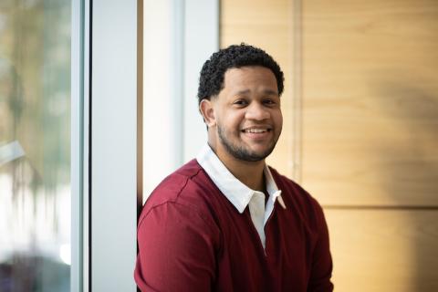 Carlos Lantigua Bari, class of 2025, is dressed in a red, long-sleeved shirt with a white collar. He's sitting near a window with wood paneling behind him. He's smiling at the camera.