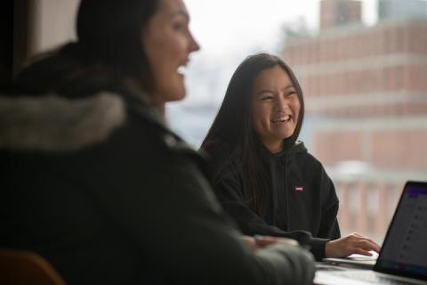 A stock photo of students studying in University Hall. Two women sit at a table in coats with their laptops and laugh while doing their work.