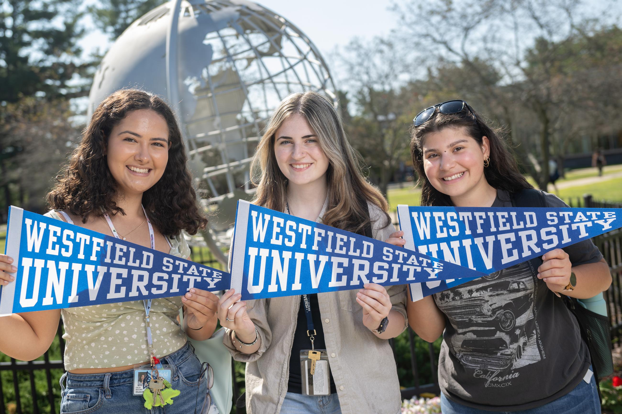 Three female Westfield State University students pose in front of the Globe holding WSU pennants.