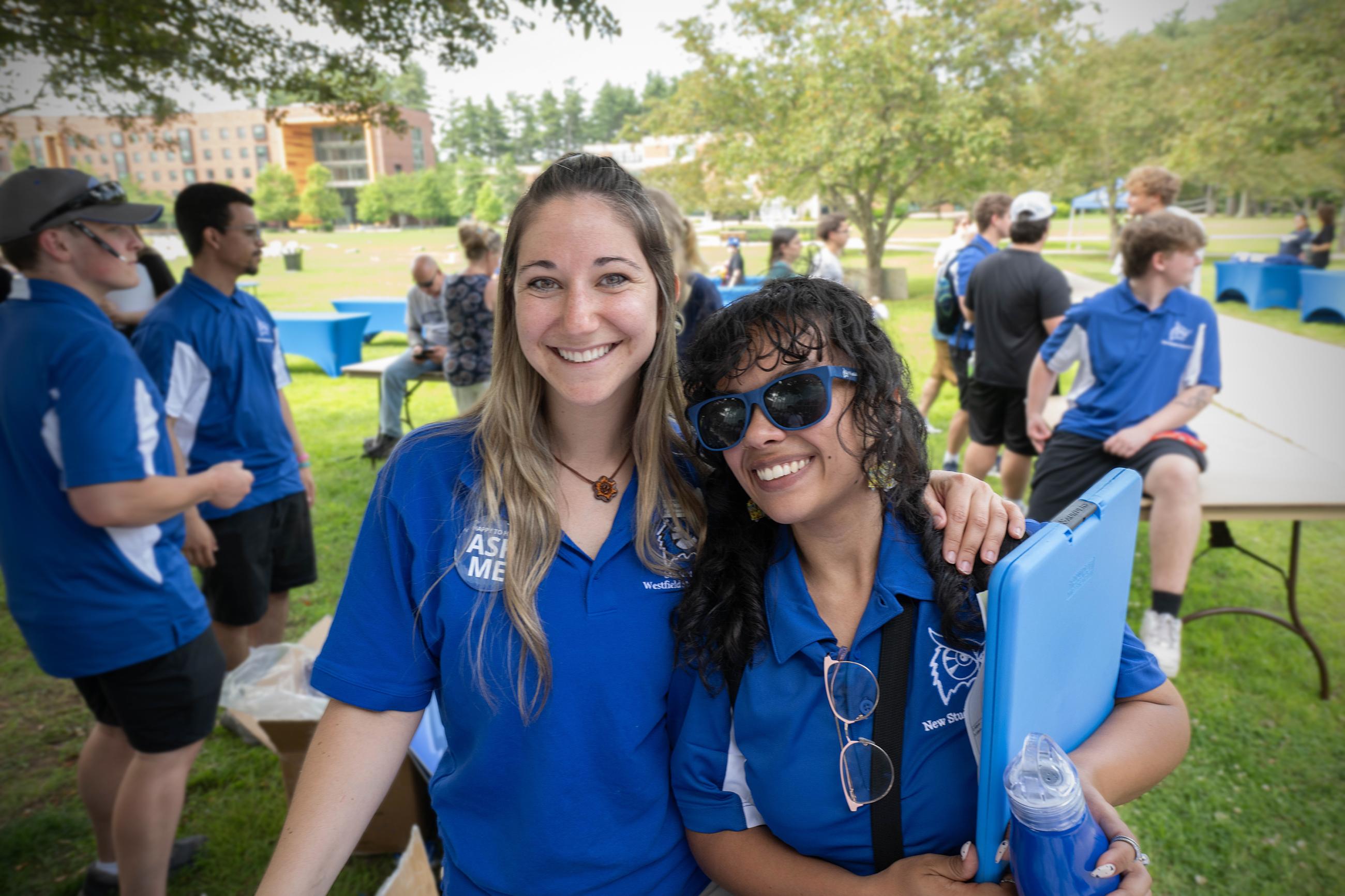 Two female Westfield State Admissions counsellors in blue shirts smile at the camera on the campus green