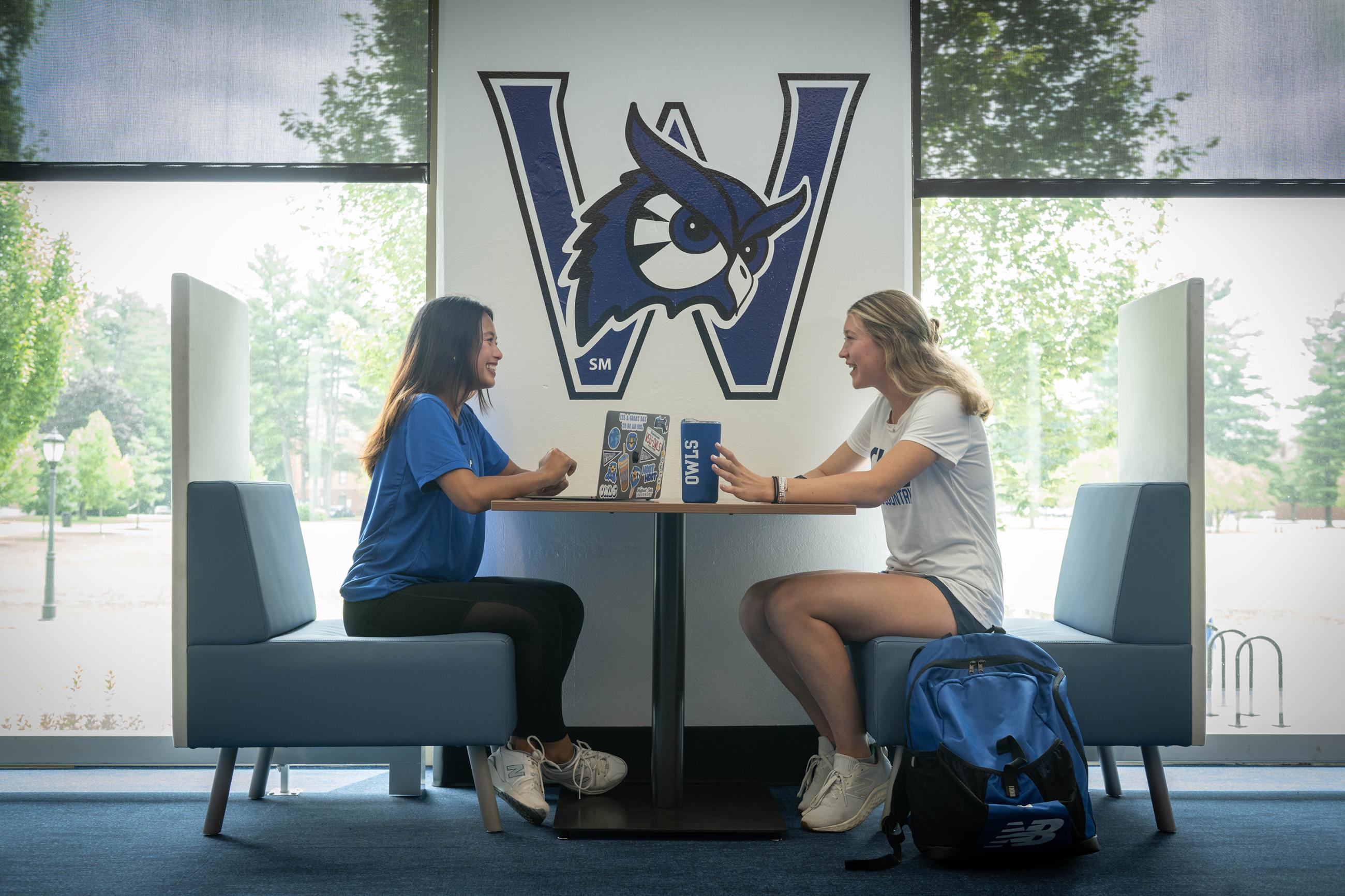 Two female students sit facing each other at a desk beside a window in the Ely Campus Center