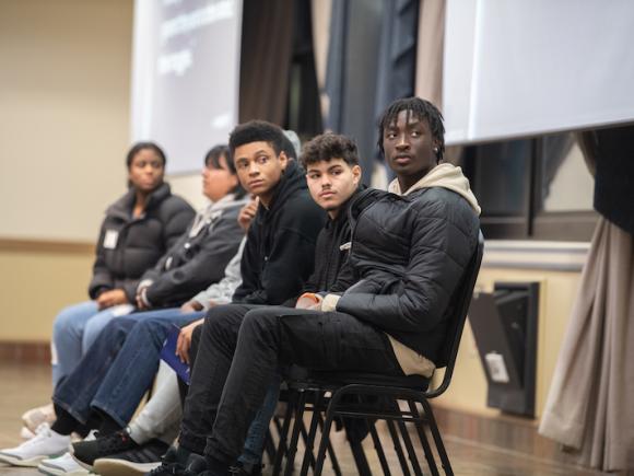 Students as part of the The Impact of Learning about Social Class in Twelfth Grade ELA on campus. They sit in a row of chairs seated next to the podium in the Scanlon Banquet Hall.