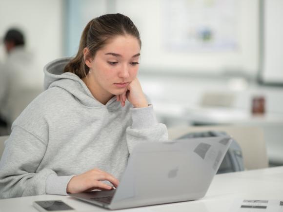 Student with laptop wearing grey sweatshirt and brown pony tail in classroom. 