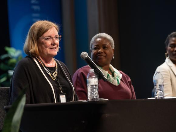 Elizabeth Dineen, a district attorney from Hampden County. She is wearing a black, long-sleeved jacket with a black undershirt. She is on stage, and speaking into a microphone as part of the "Supporting Our Superwomen" symposium.