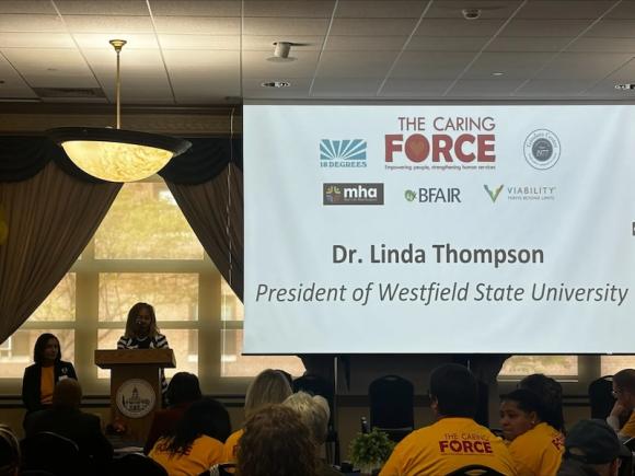 President Linda Thompson speaking at the The Western MA Caring Force Rally, held on September 17 at Westfield State University’s Scanlon Hall. She is standing at a podium and speaking on behalf of the University in front of people in yellow shirts, with a projector screen next to her.