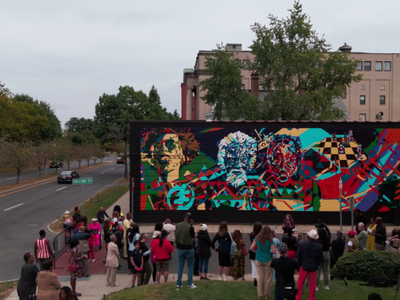 A crowd gathers outside a mural by AfriCOBRA raist and muralist Nelson Stevens in the documentary "Art for the People," directed by Evan Goodchild. Credits to Evan Goodchild and Goodchild Media.