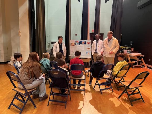 Westfield State Physician Assistant students teach younger students from Westfield Intermediate School. Three PA students in white lab coats present a poster to several younger students who sit in chairs before them.