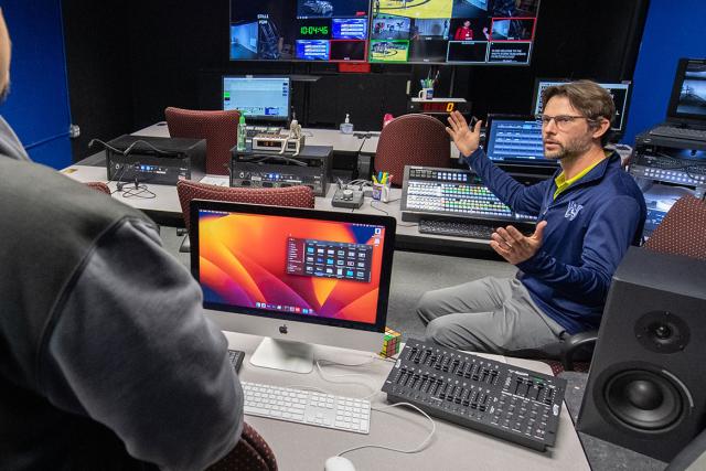 A professor seated in a television production studio instructs their students.