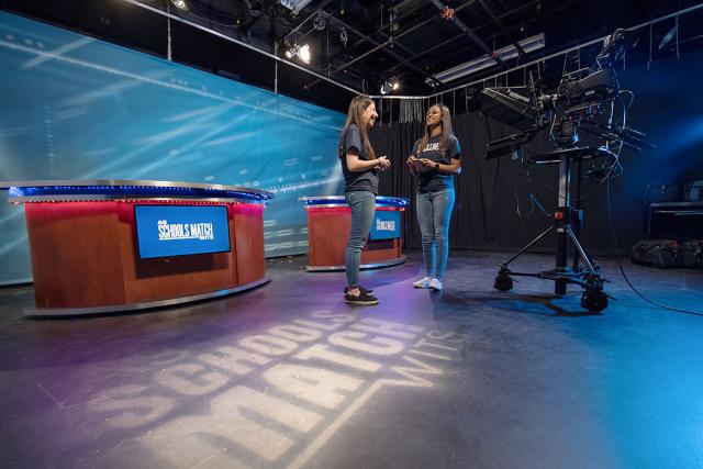 Two Westfield State University students smile as they talk to each other in a news studio.