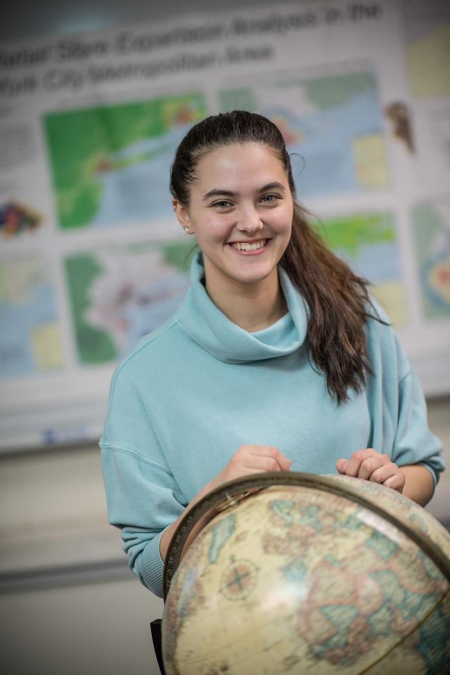 Student smiling wearing blue sweater in front of a globe.