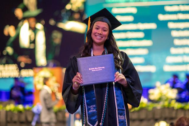 Student smiling holding diploma proudly at commencement ceremony.