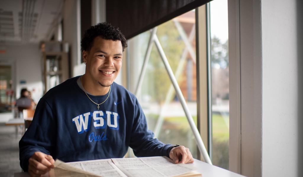 TRIO student wearing blue WSU shirt smiling with book open.