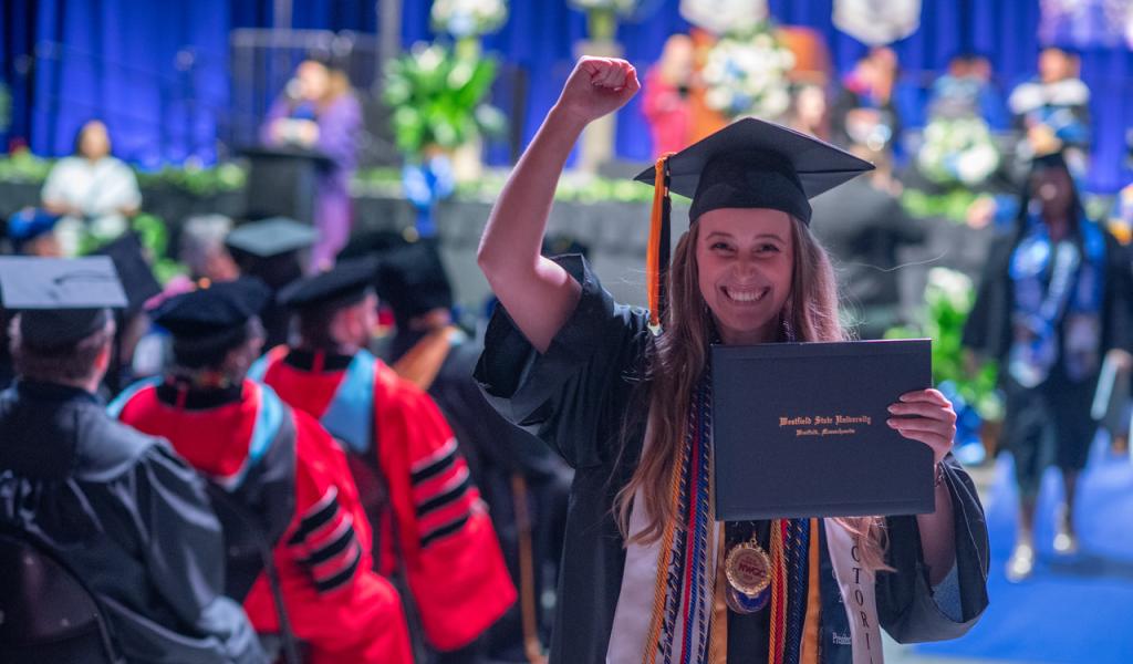 Student wearing cap and gown at commencement holding diploma smiling.