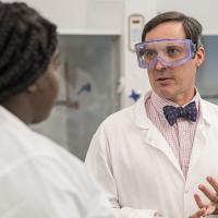A professor wearing a lab coat and safety glasses talks to a student in a science lab.