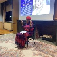 Woman sits in front of presentation screen and speaks to an audience